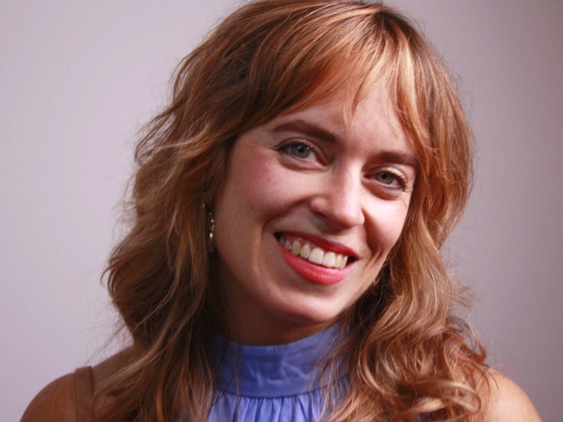 Professional portrait of Nicole Biros-Bolton, employment and human rights lawyer, smiling warmly against a neutral background. She has wavy light brown hair and is wearing a blue blouse.