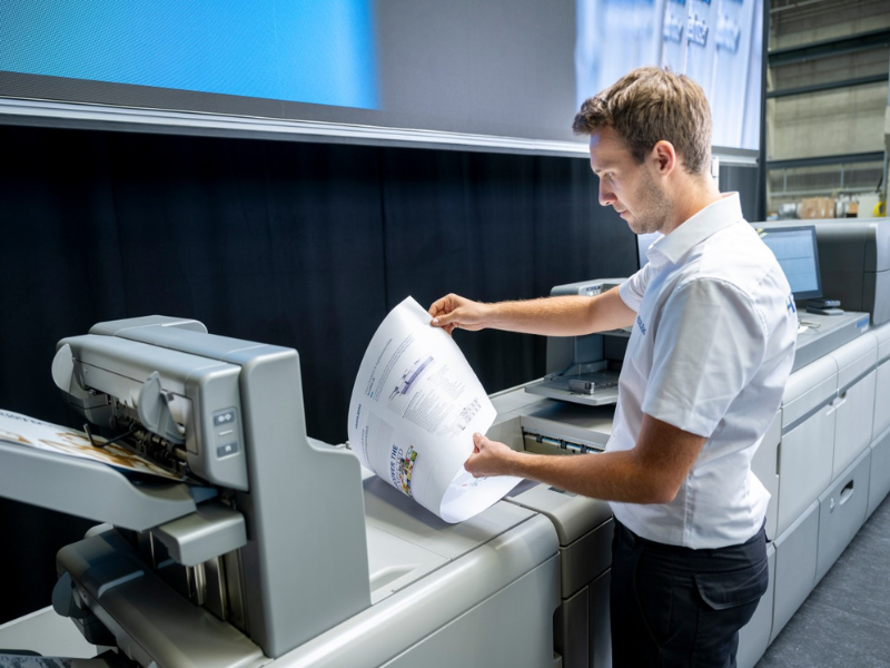 Digital Press Investment as Print Strategy Print operator reviewing a printed proof beside a Heidelberg digital press during a production quality check.